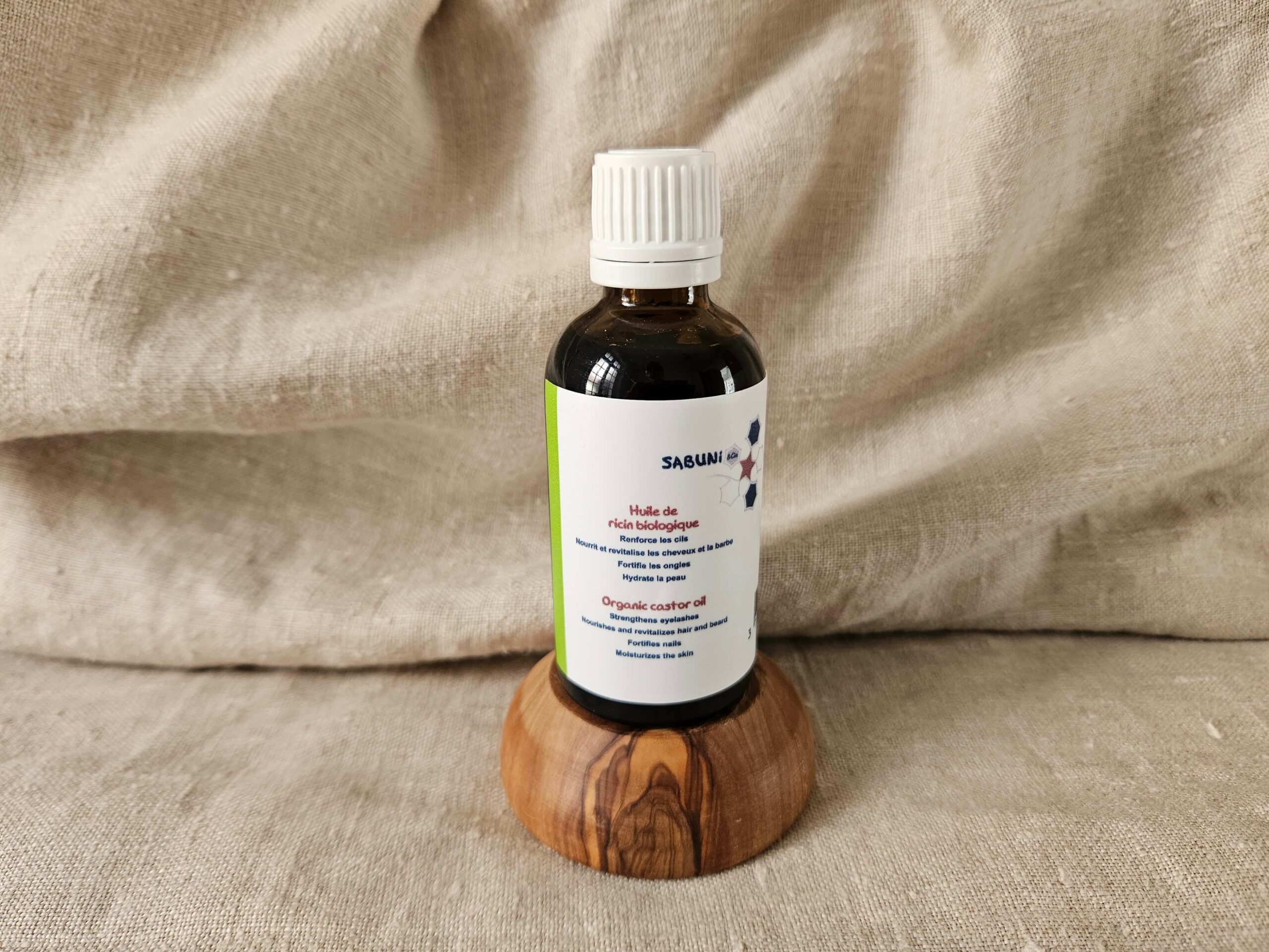 Huile de Ricin biologique, reconditionnée par la savonnerie artisanale Sabuni & Cie, dans un flacon en verre opaque, posé sur un socle en olivier.