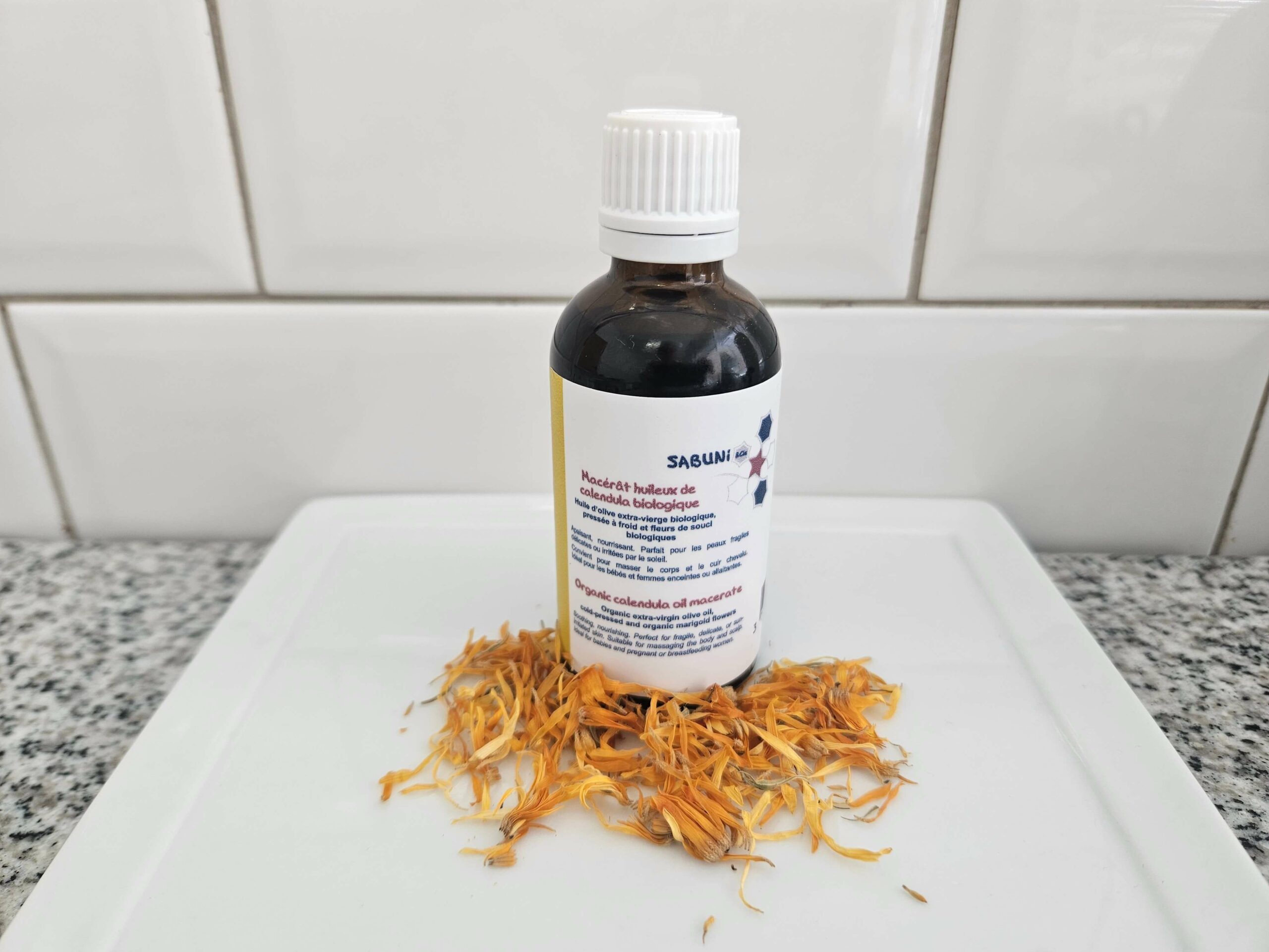 Macérat huileux de calendula précieux, fait à la main, par la savonnerie artisanale Sabuni & Cie, selon la recette ancestrale, dans un flacon en verre opaque, posé devant du carrelage style métro.