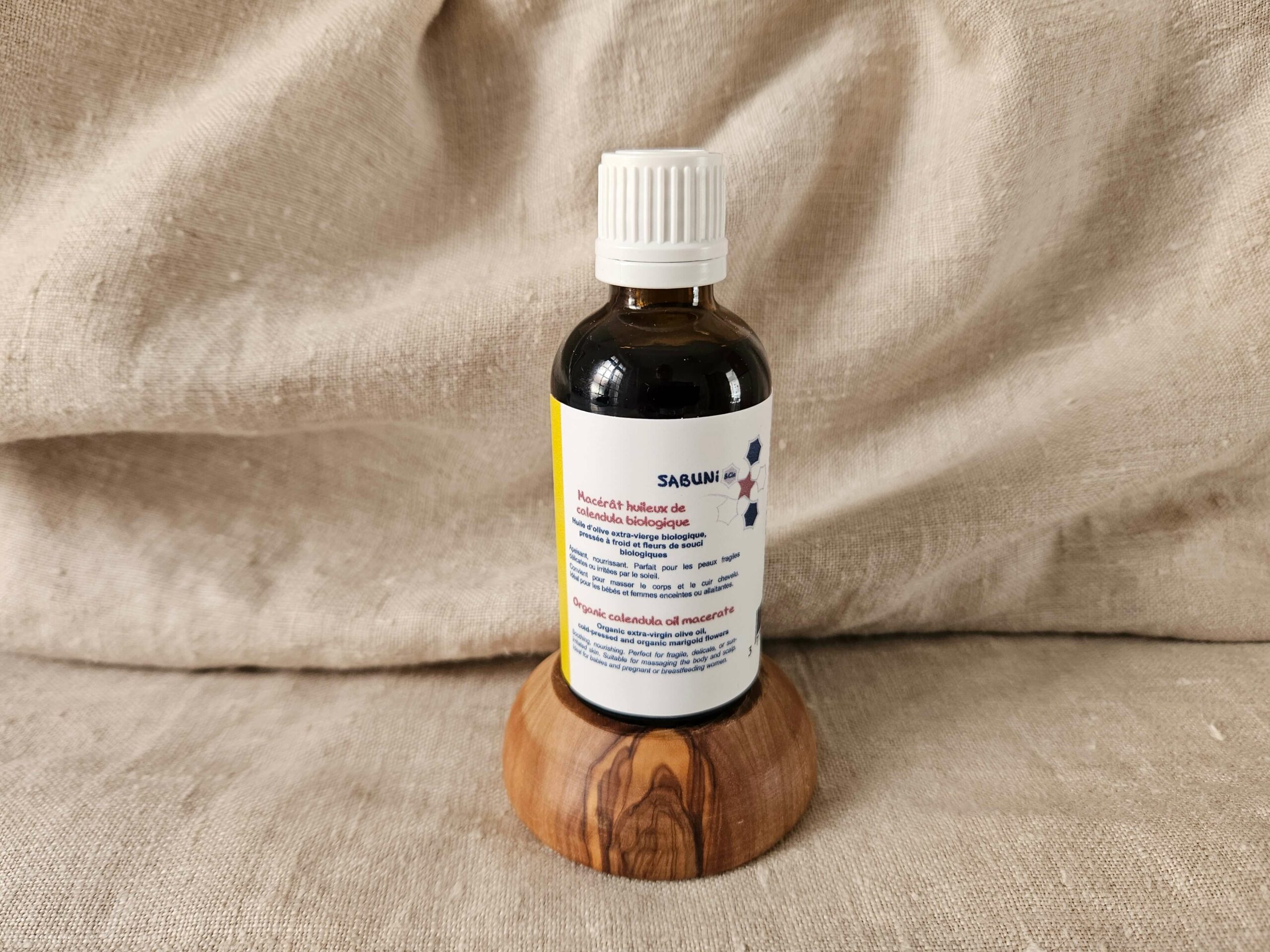 Macérat huileux de calendula précieux, fait à la main, par la savonnerie artisanale Sabuni & Cie, selon la recette ancestrale, dans un flacon en verre opaque, posé sur un socle en olivier.